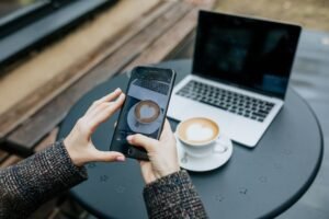 Lady's hands holding a smartphone and taking a photo of her coffee at a cafe table with laptop in background