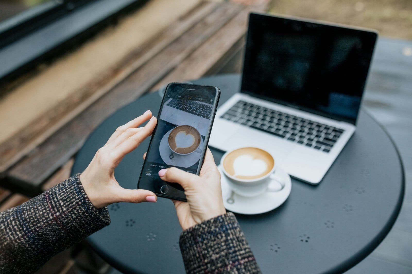 Lady's hands holding a smartphone and taking a photo of her coffee at a cafe table with laptop in background